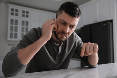Photo of Emotional man talking on phone at table in kitchen. Hate concept