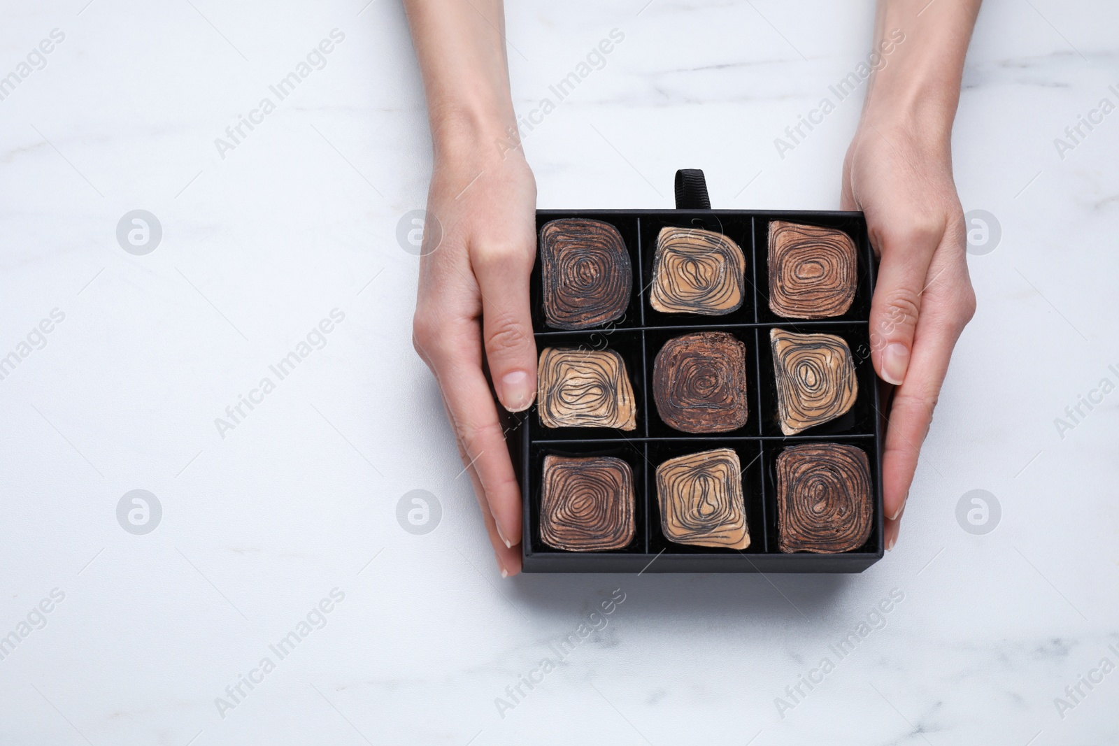 Photo of Woman holding box of delicious chocolate candies on white marble table, top view. Space for text