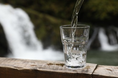 Photo of Fresh water pouring into glass on wooden surface near waterfall. Space for text