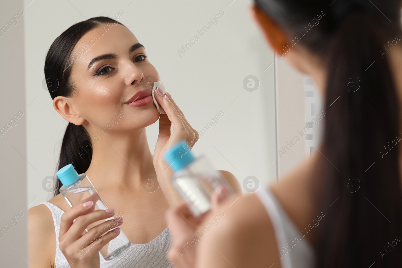 Photo of Beautiful woman removing makeup with cotton pad near mirror indoors