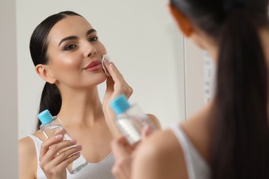 Beautiful woman removing makeup with cotton pad near mirror indoors