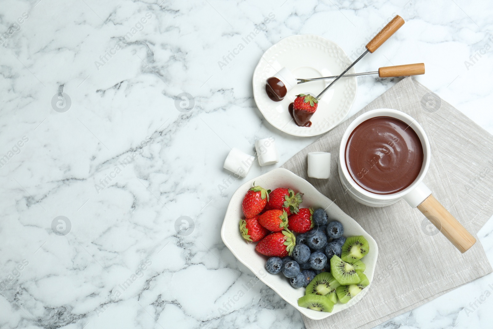Photo of Fondue pot with melted chocolate, sweet marshmallows, fresh kiwi, different berries and forks on white marble table, flat lay. Space for text