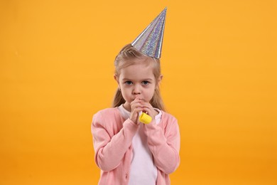 Birthday celebration. Cute little girl in party hat with blower on orange background