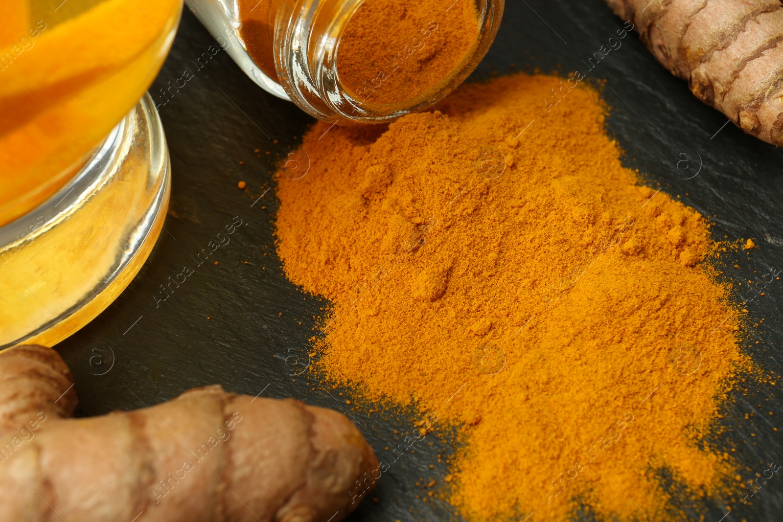 Photo of Glass cup of tasty tea, turmeric roots and powder on black textured table, closeup