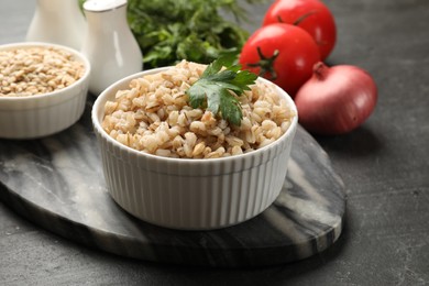 Photo of Tasty pearl barley porridge and parsley in bowl on dark textured table, closeup