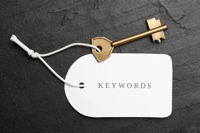 Photo of Metal key and tag wIth word KEYWORDS on black table, top view