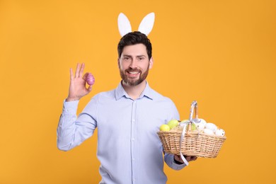 Portrait of happy man in cute bunny ears headband holding wicker basket with Easter eggs on orange background