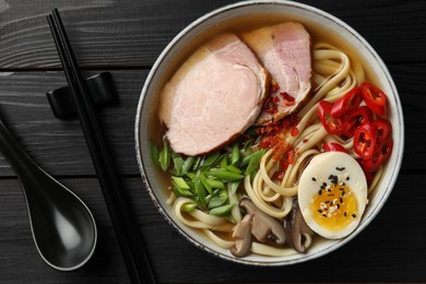 Delicious ramen in bowl, spoon and chopsticks on black wooden table, flat lay. Noodle soup