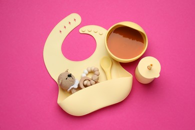Photo of Silicone baby bib, toys and plastic dishware with healthy food on pink background, flat lay