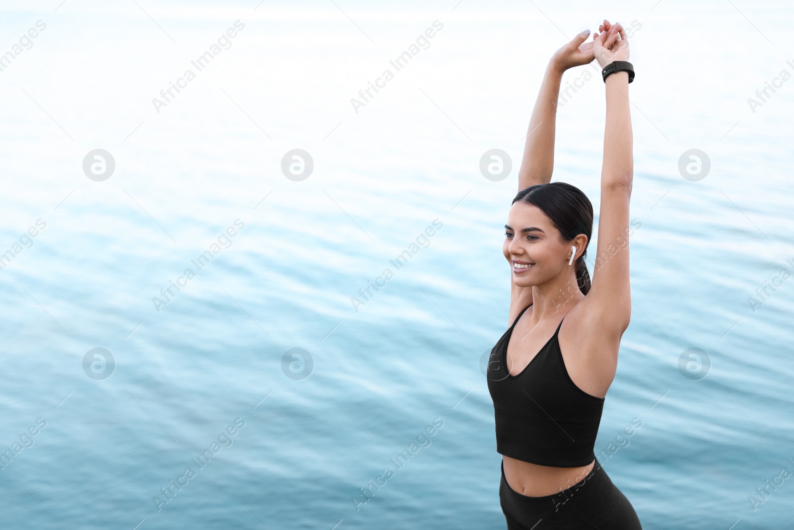 Photo of Young sportswoman with wireless earphones stretching near river