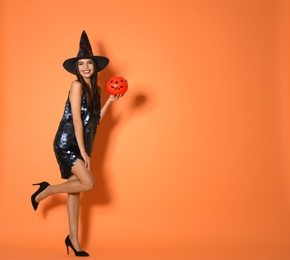 Photo of Beautiful woman wearing witch costume with Jack O'Lantern candy container for Halloween party on yellow background, space for text