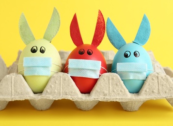 Photo of Dyed eggs with bunny ears and protective masks in carton on yellow background, closeup. Easter holiday during COVID-19 quarantine