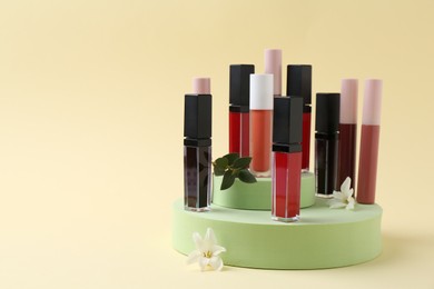 Photo of Different lip glosses, podiums and flowers on beige background, space for text