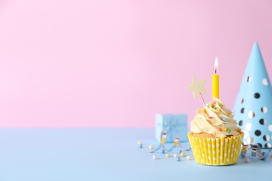 Delicious birthday cupcake with burning candle, party decor and gift box on light blue table against pink background, space for text