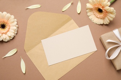 Image of Beautiful herbera flowers, gift, envelope and blank card with space for text on brown background, flat lay