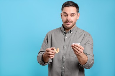 Photo of Handsome man holding tasty fortune cookie and reading prediction on light blue background. Space for text