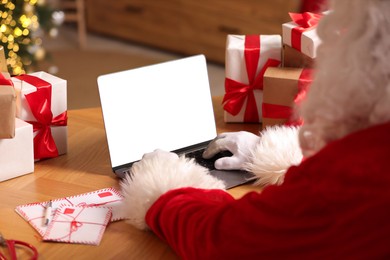 Photo of Santa Claus using laptop, closeup. Christmas gifts and letters on table indoors