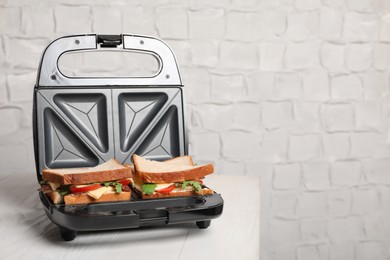 Photo of Modern grill maker with tasty sandwiches and ingredients on white wooden table. Space for text