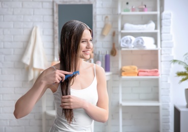 Photo of Young woman brushing hair after applying mask in bathroom