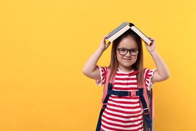 Photo of Cute little girl in glasses with open book and backpack against orange background. Space for text