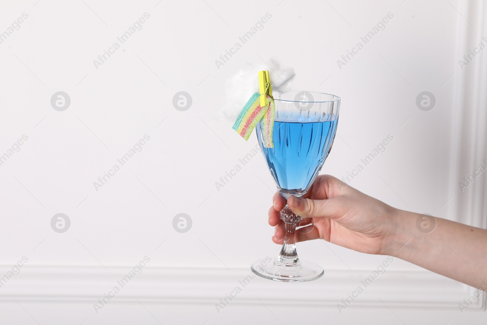 Photo of Woman holding glass of cocktail decorated with cotton candy and sour rainbow belt near white wall, closeup. Space for text