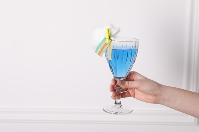 Photo of Woman holding glass of cocktail decorated with cotton candy and sour rainbow belt near white wall, closeup. Space for text