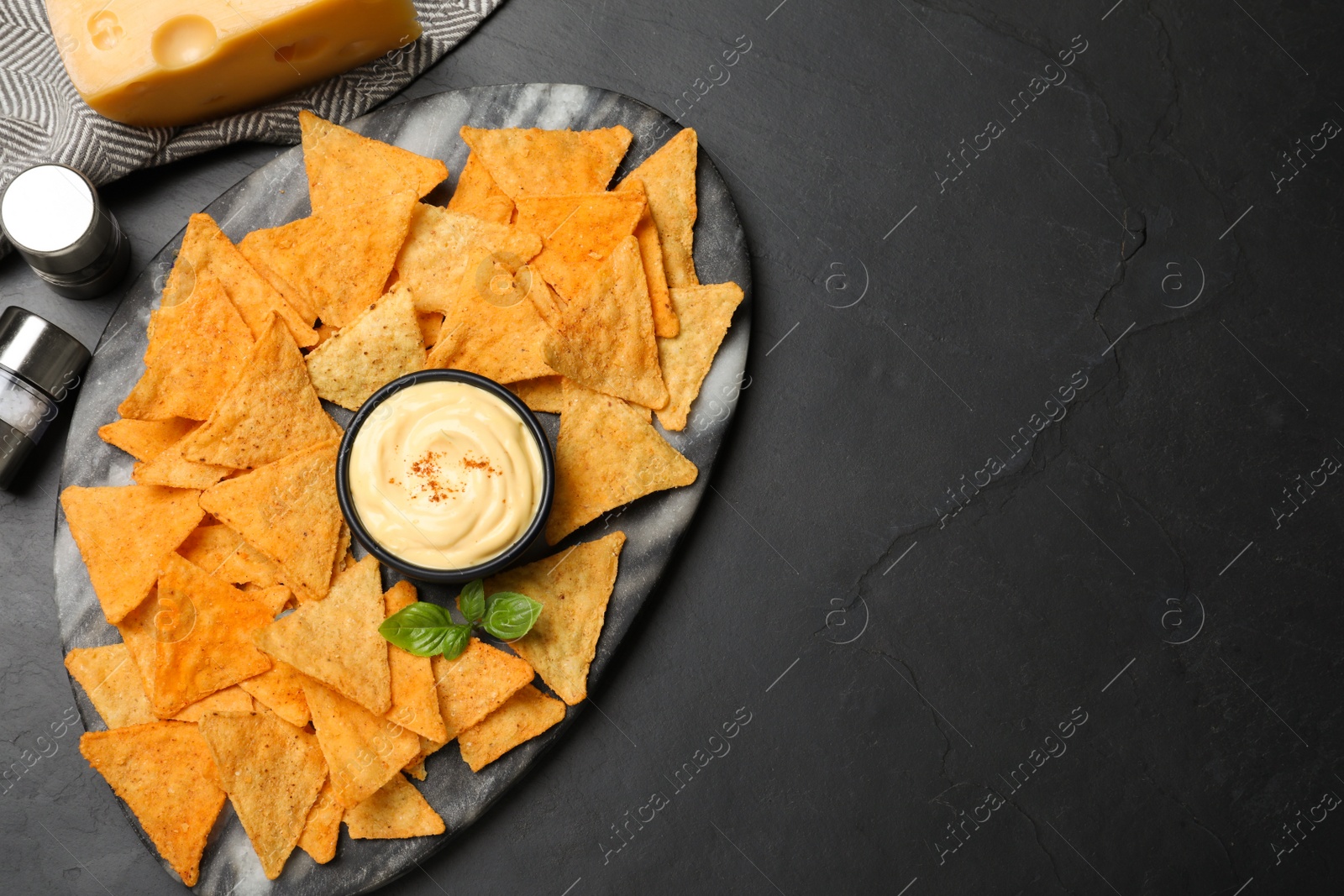 Photo of Delicious nachos and cheese sauce with basil on black table, flat lay. Space for text