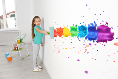 Little girl drawing rainbow on white wall indoors