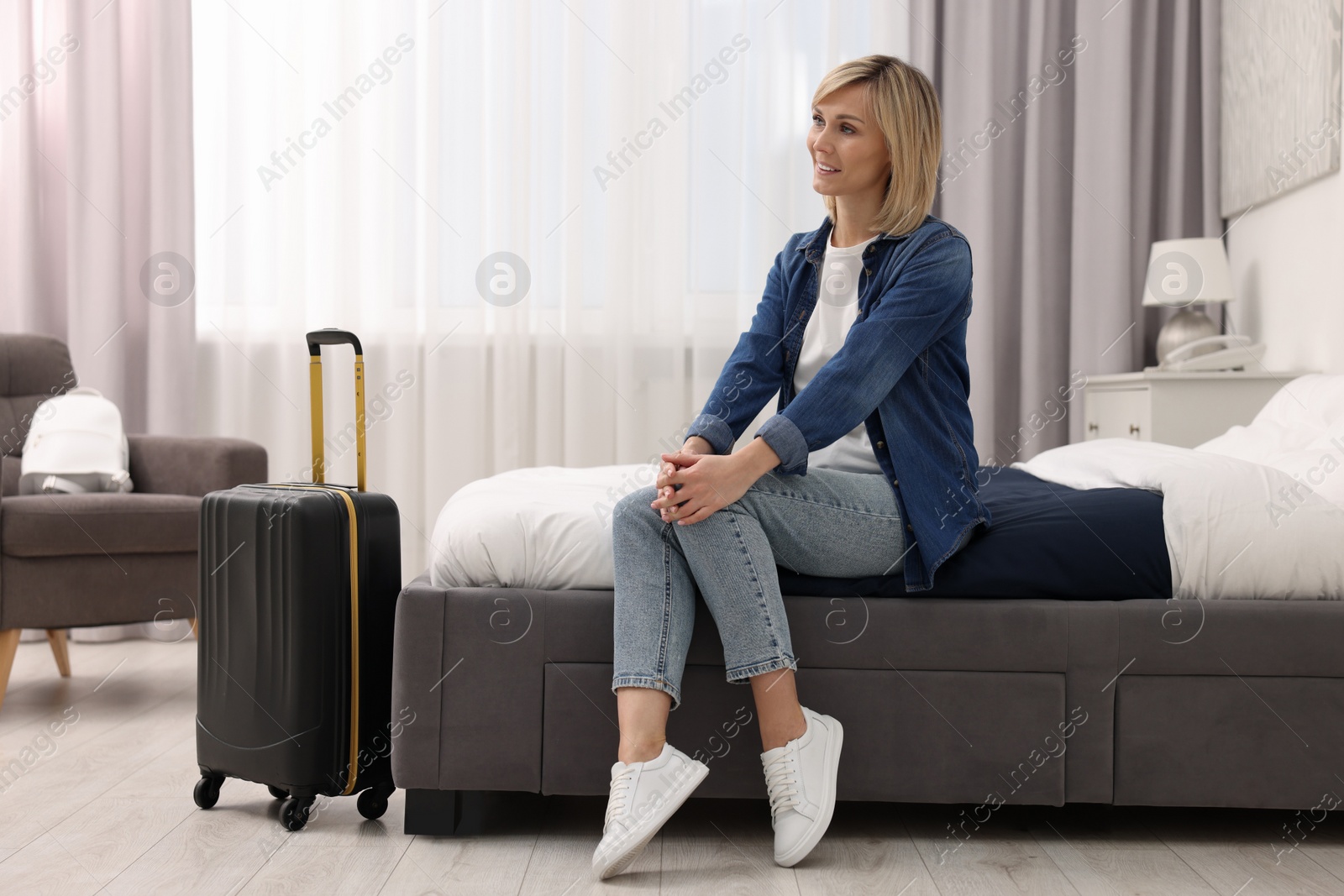 Photo of Smiling guest with suitcase relaxing on bed in stylish hotel room