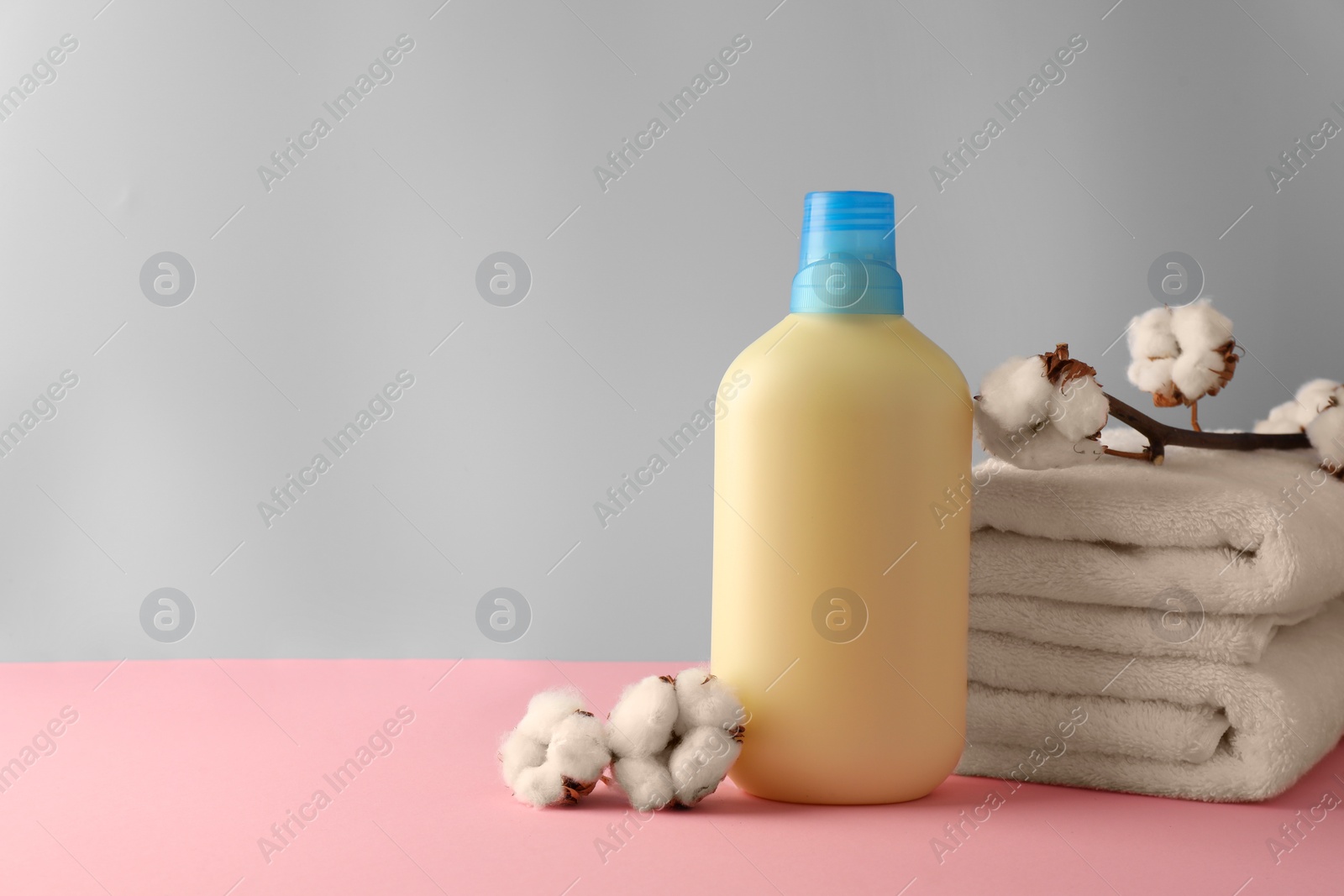 Photo of Bottle of fabric softener, stacked clean towels and fluffy cotton flowers on pink table against light grey background, space for text