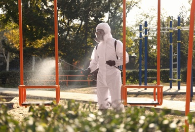 Photo of Man in hazmat suit spraying disinfectant onto swing at children's playground. Surface treatment during coronavirus pandemic