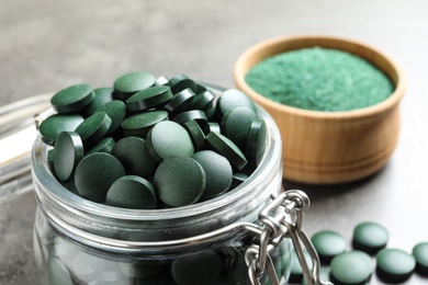 Photo of Glass jar with green spirulina pills on grey table, closeup