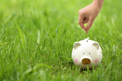 Photo of Young woman putting coin into piggy bank on green grass outdoors, closeup. Space for text