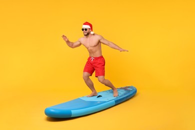 Photo of Happy man in Santa hat balancing on SUP board against orange background