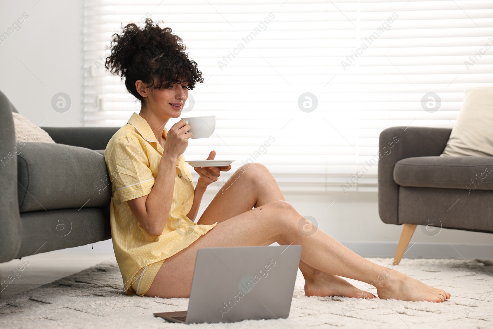 Photo of Beautiful young woman in stylish pyjama with cup of drink and laptop on floor at home