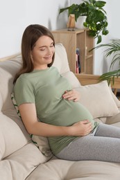 Photo of Happy pregnant woman sitting on sofa and touching her belly in living room