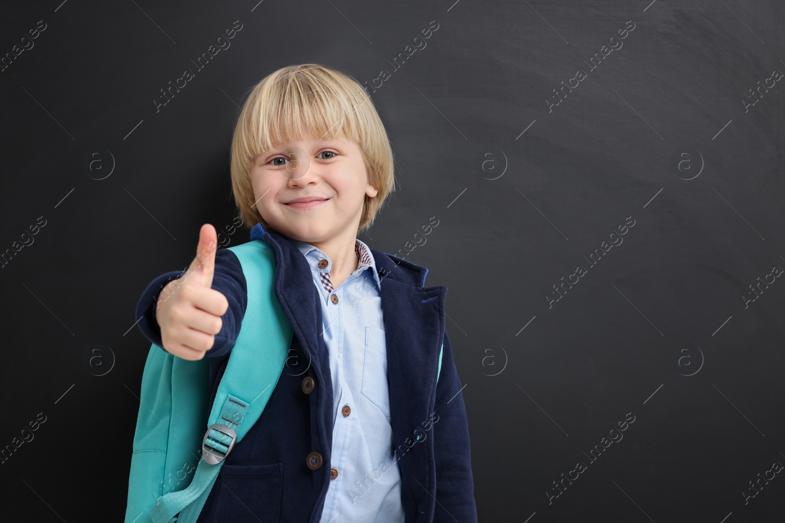 Photo of Happy little school child with backpack showing thumbs up near chalkboard. Space for text