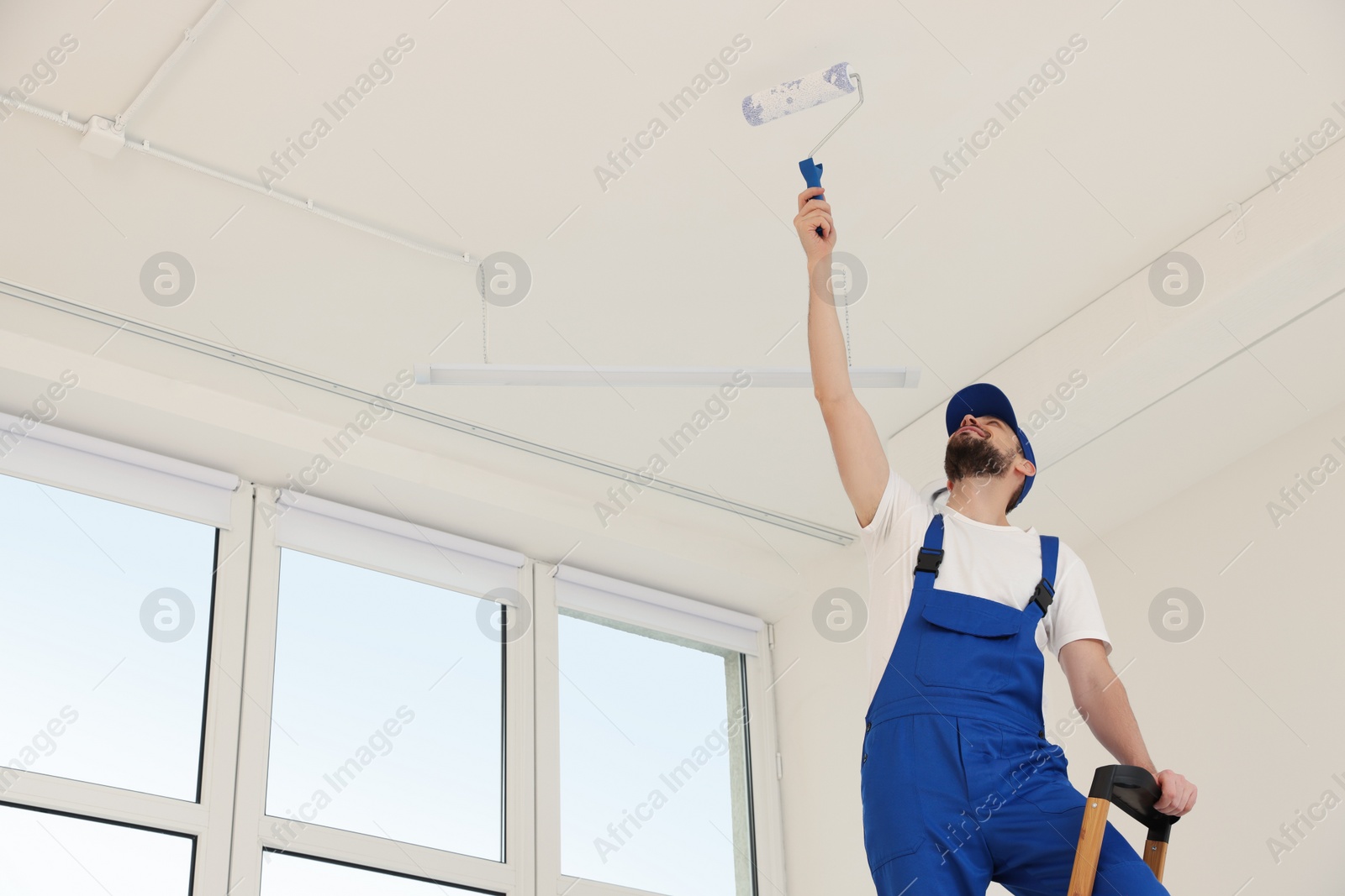 Photo of Worker in uniform painting ceiling with roller indoors, low angle view