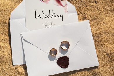Photo of Envelopes with wedding invitations and gold rings on sandy beach, top view