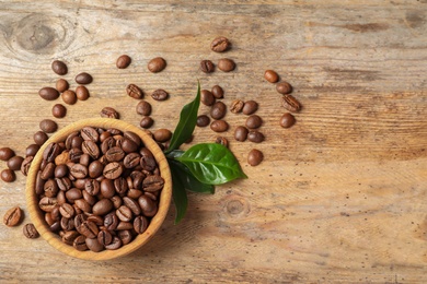 Bowl of coffee beans and fresh green leaves on wooden table, top view with space for text