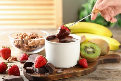 Photo of Woman dipping strawberry into fondue pot with chocolate at wooden table, closeup