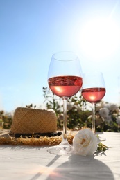 Glasses of rose wine, straw hat and beautiful flower on white wooden table outdoors