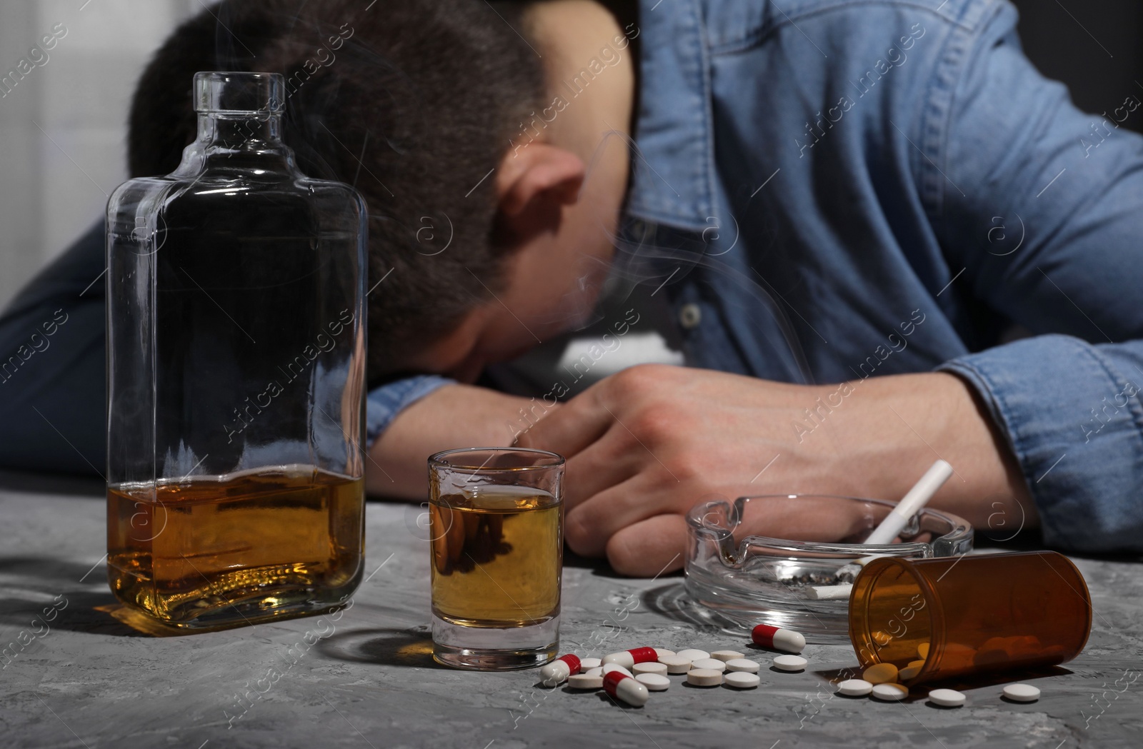 Photo of Alcohol and drug addiction. Man with whiskey, smoldering cigarettes and pills at grey textured table, selective focus