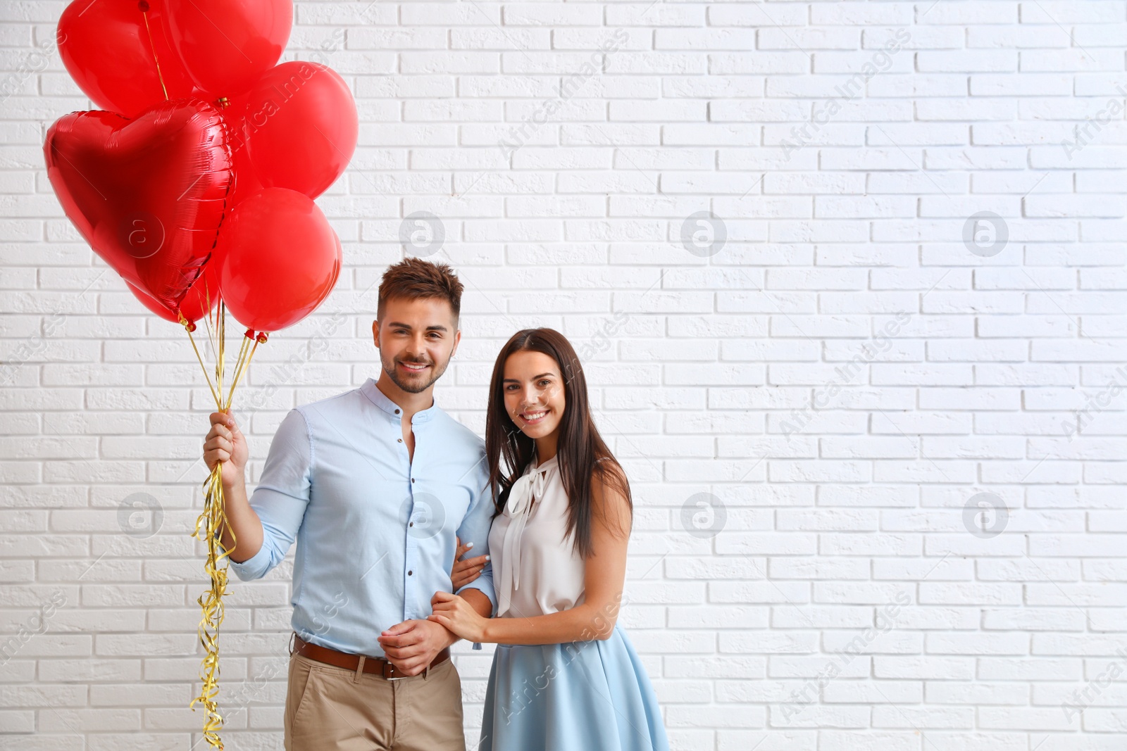 Photo of Young couple with air balloons near white brick wall. Celebration of Saint Valentine's Day