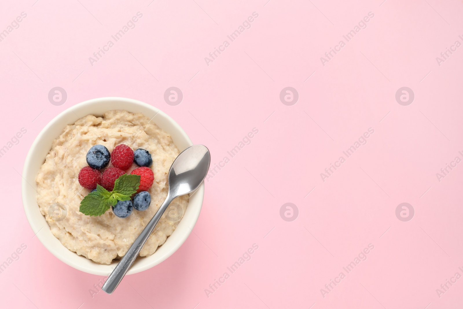 Photo of Tasty oatmeal porridge with raspberries, blueberries and spoon in bowl on pink background, top view