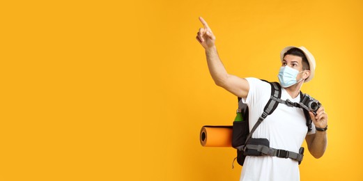 Male tourist in protective mask with travel backpack and camera on yellow background, space for text