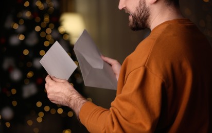 Photo of Man holding envelope and greeting card against blurred Christmas lights, closeup