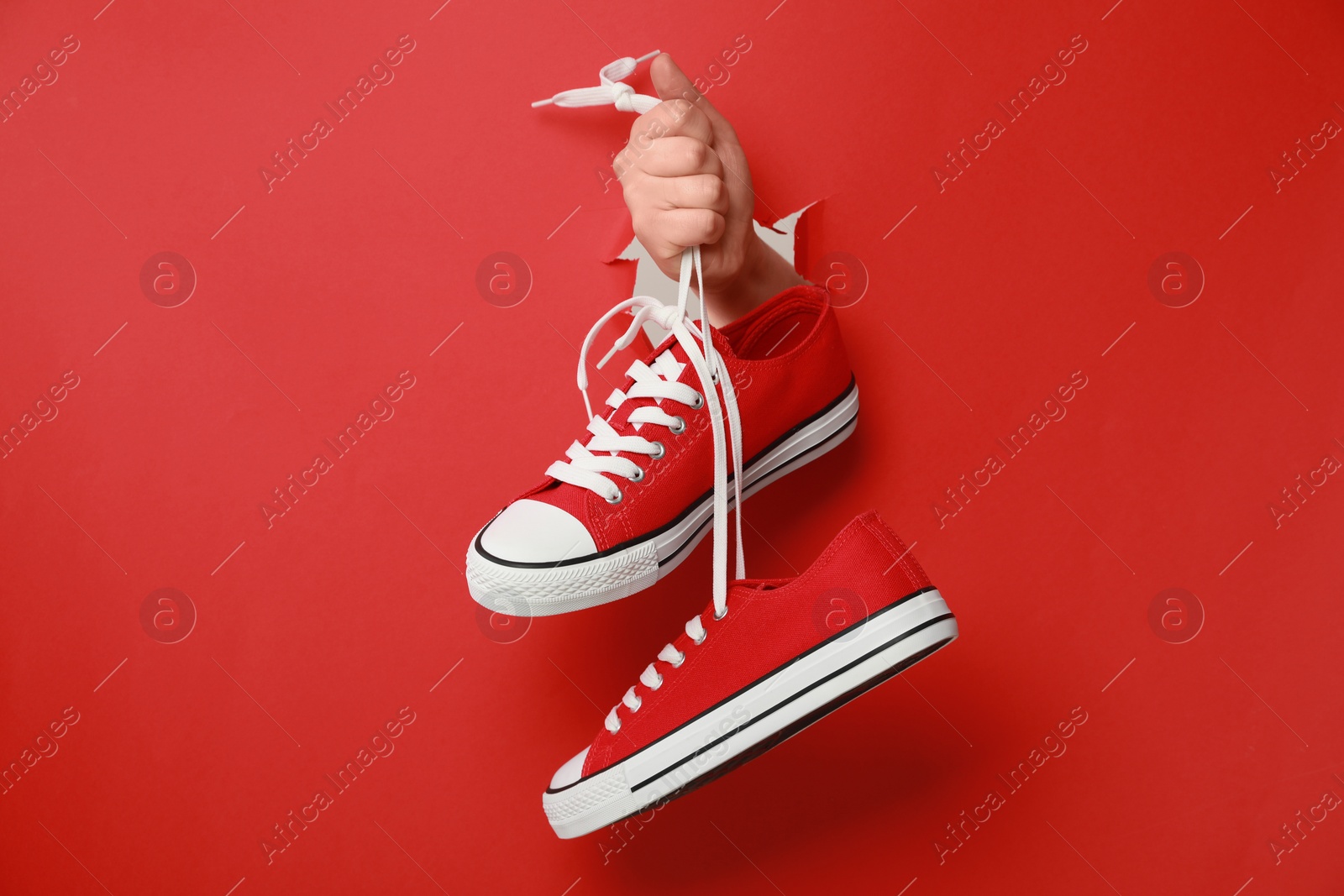 Photo of Woman holding pair of classic old school sneakers through hole in red paper, closeup