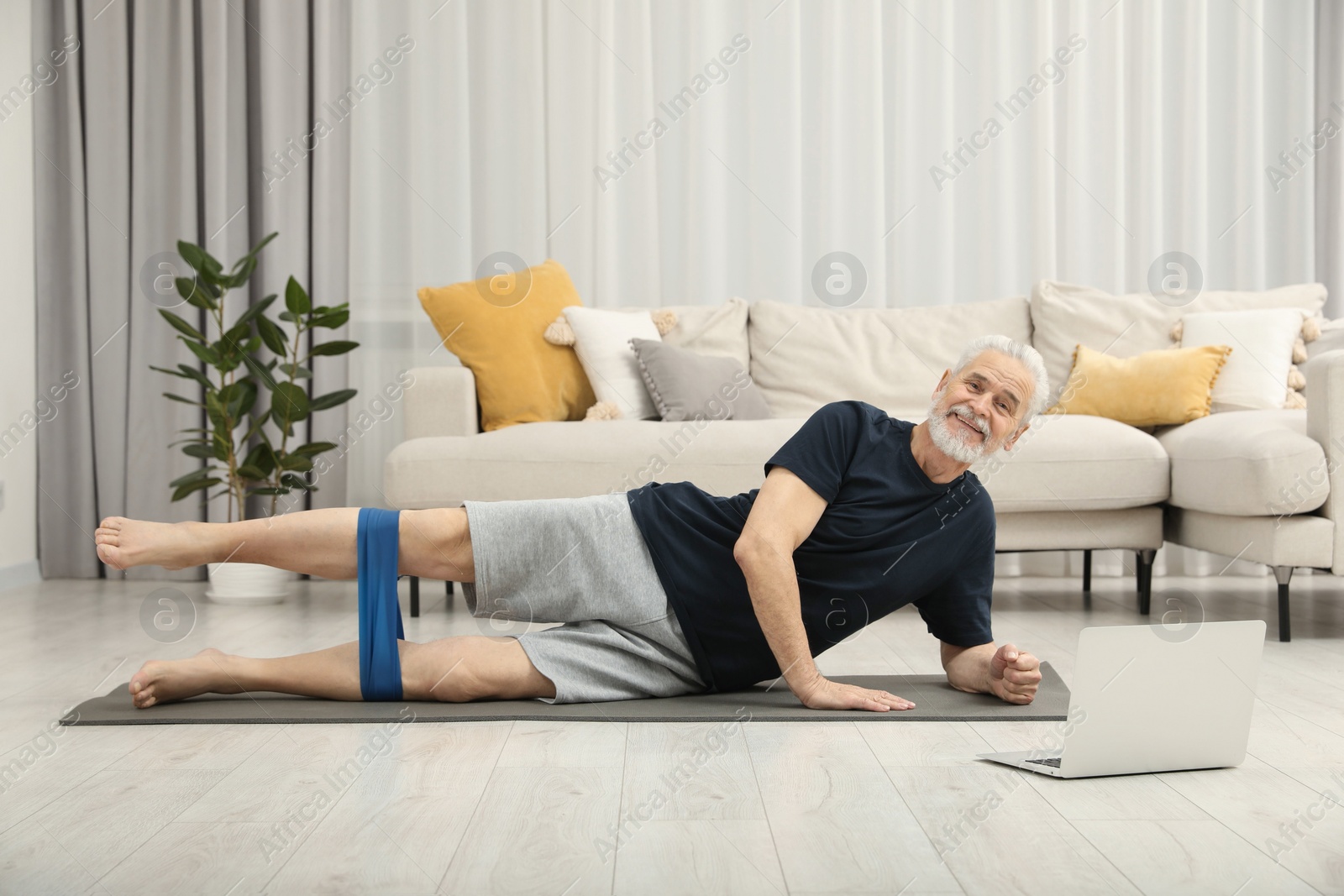 Photo of Senior man doing exercise with fitness elastic band near laptop on mat at home