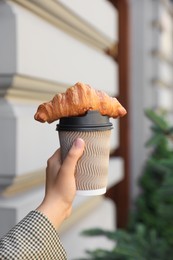 Photo of Woman holding tasty croissant and cup of coffee outdoors, closeup